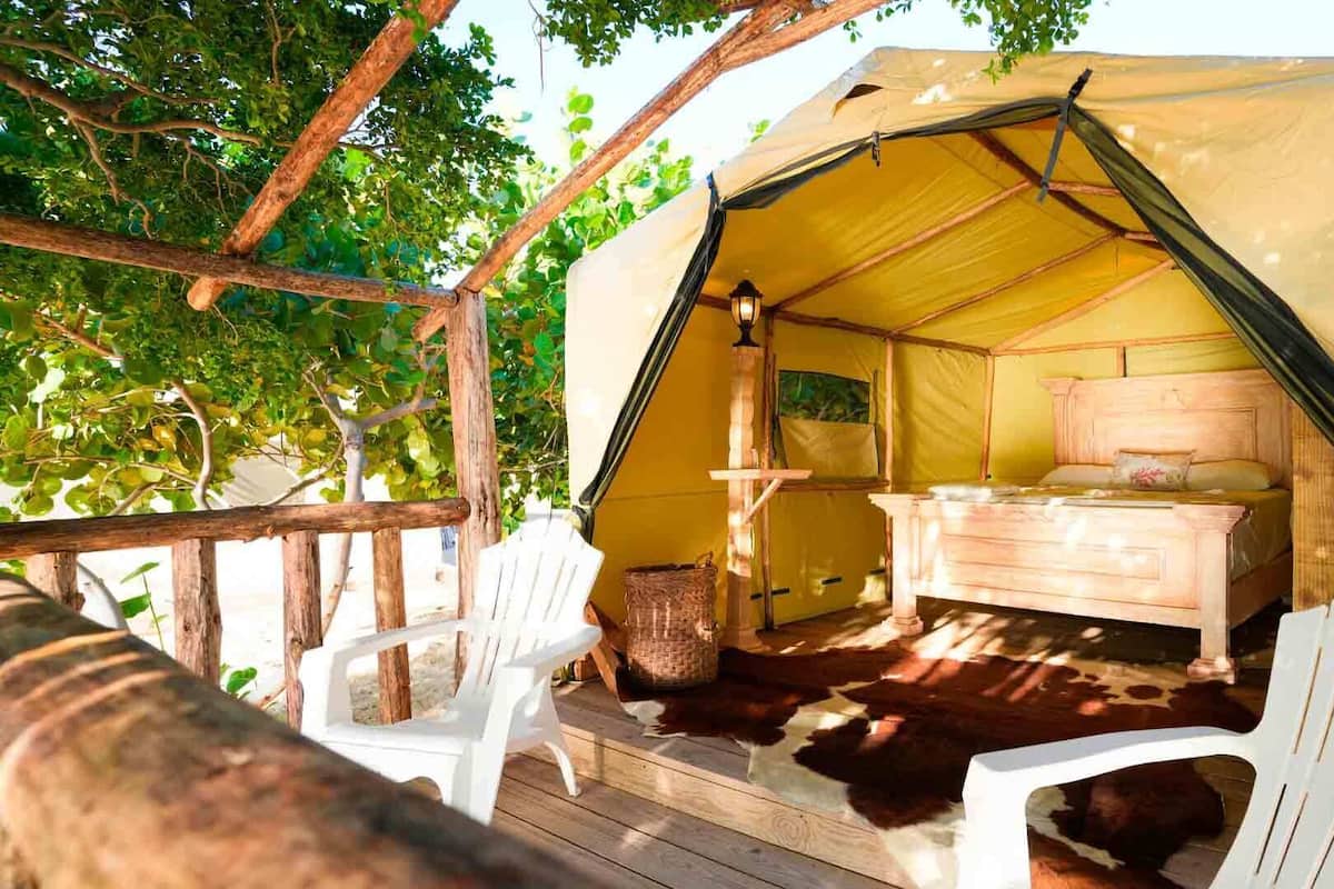 Une tente avec un lit en bois et deux chaises blanches, installée sur une terrasse en bois recouverte d’un tapis en peau de vache.