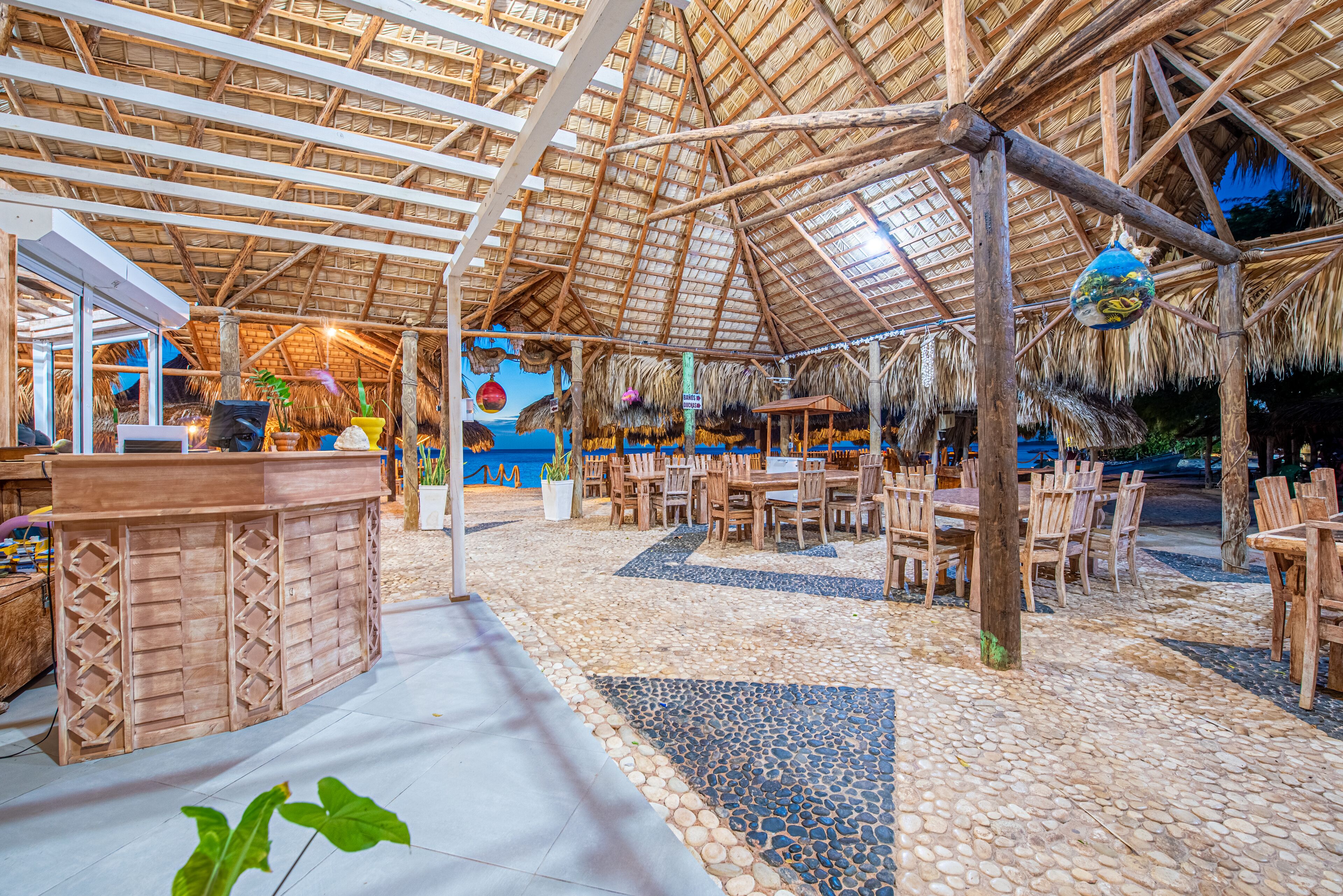 Espaço de refeições ao ar livre com teto de palha, mesas e cadeiras de madeira, e um bar central.