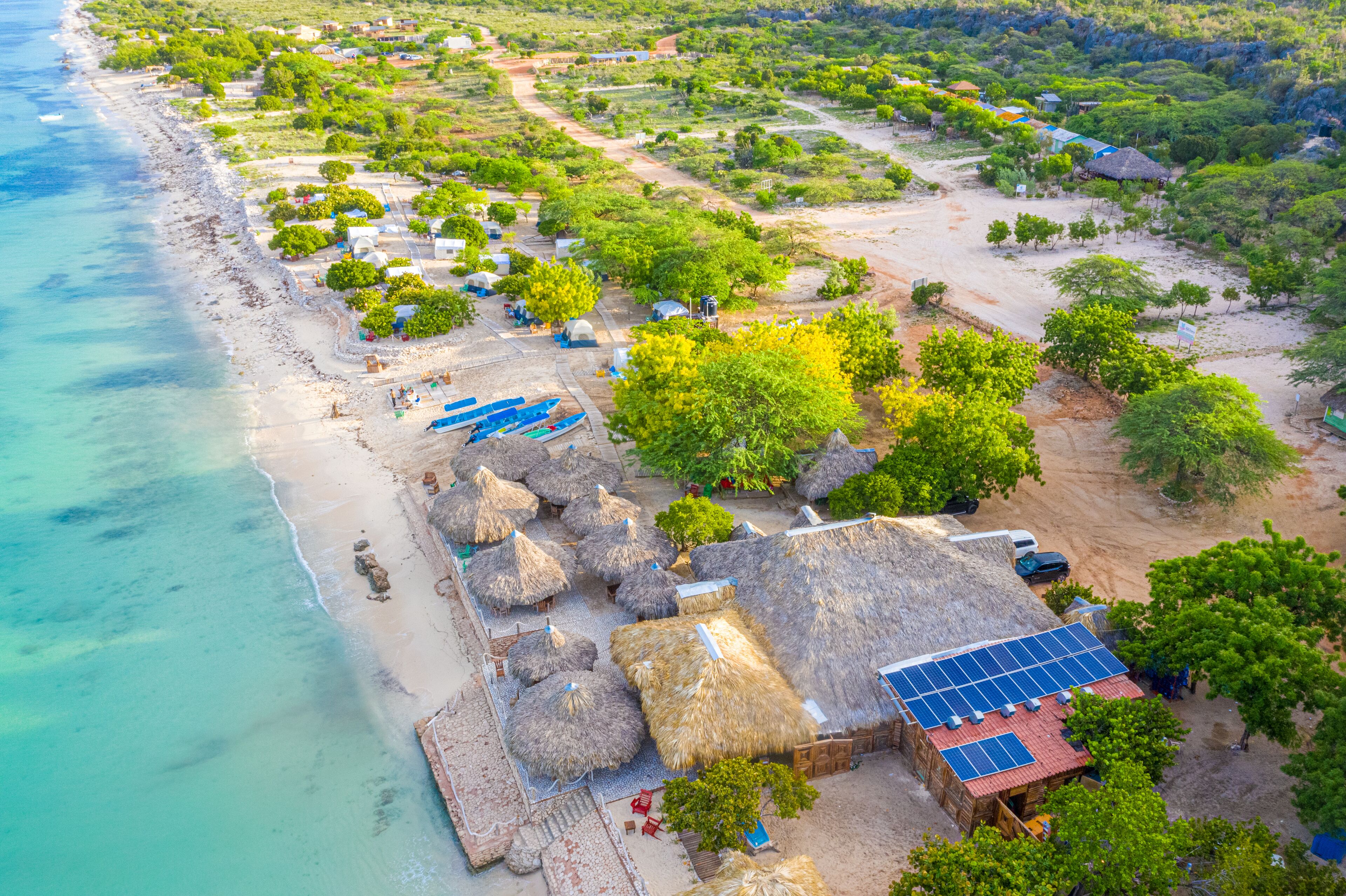 An aerial view of a beachside resort with thatched-roof huts, solar panels, and lush greenery.