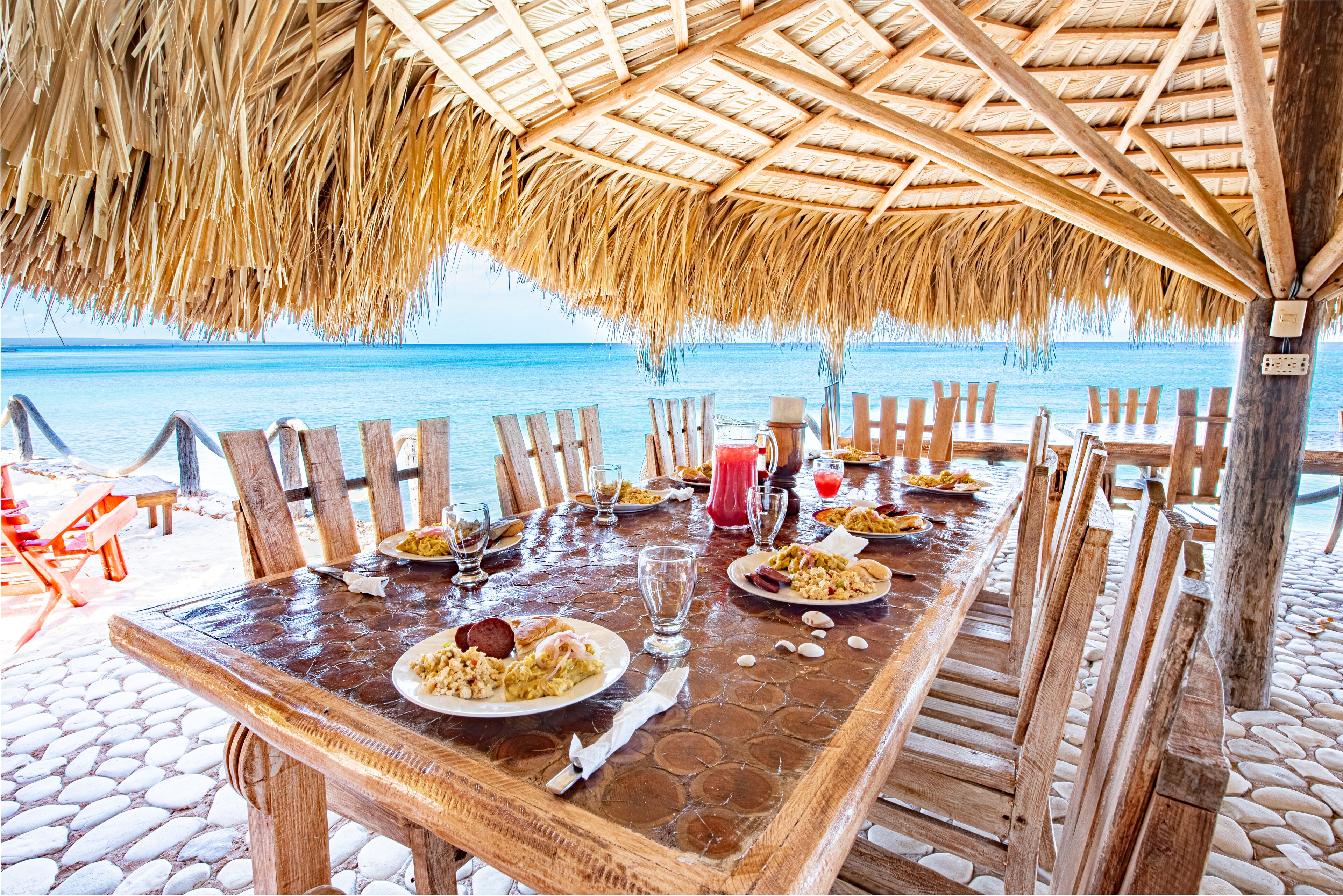 A wooden table set with plates of food, glasses, and a drink, under a thatched roof with a view of the ocean.