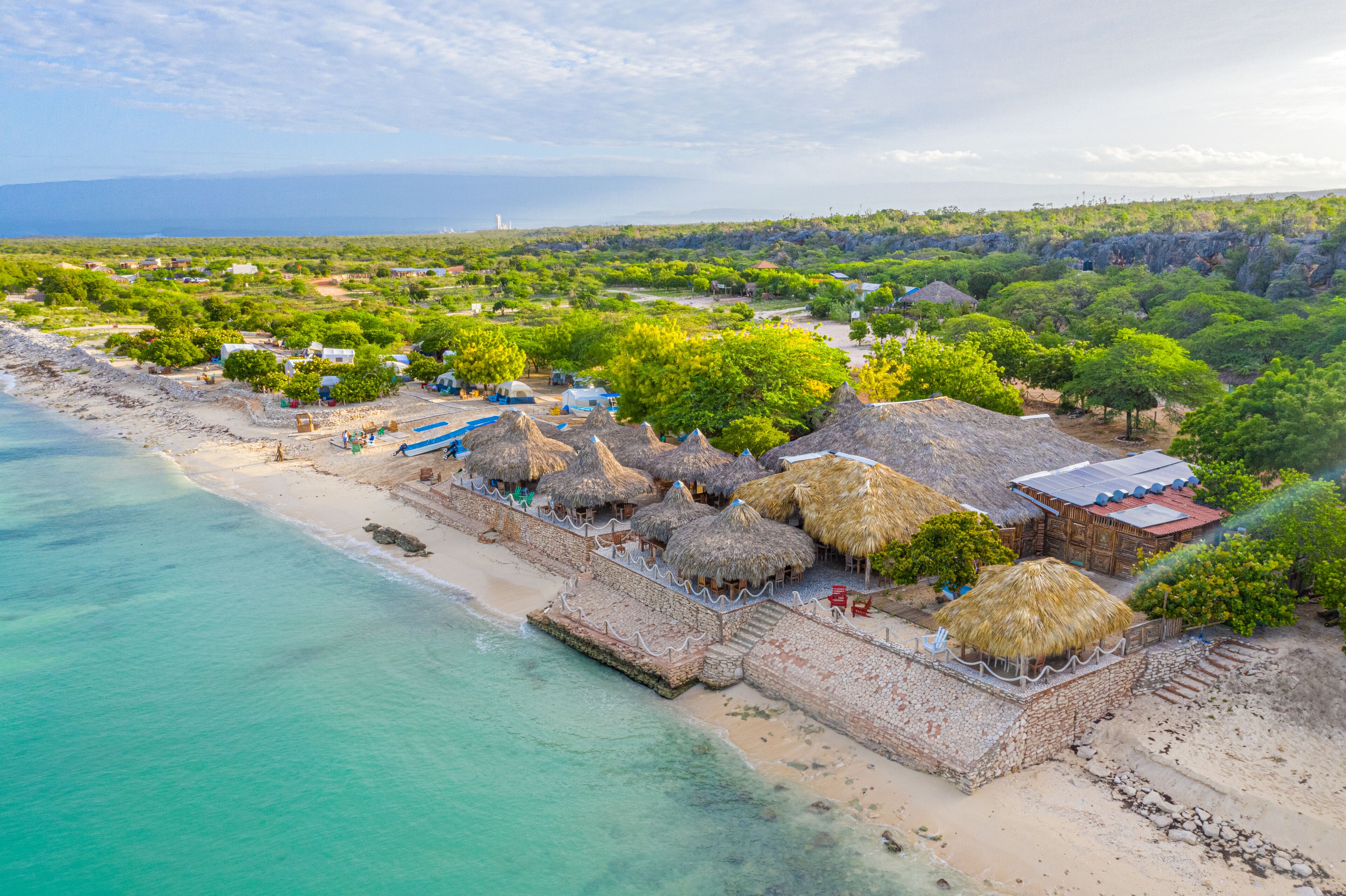 An aerial view of a beach resort with thatched-roof huts, a sandy beach, and a clear view of the ocean.