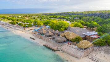 An aerial view of a beach resort with thatched-roof huts, a sandy beach, and a clear view of the ocean.