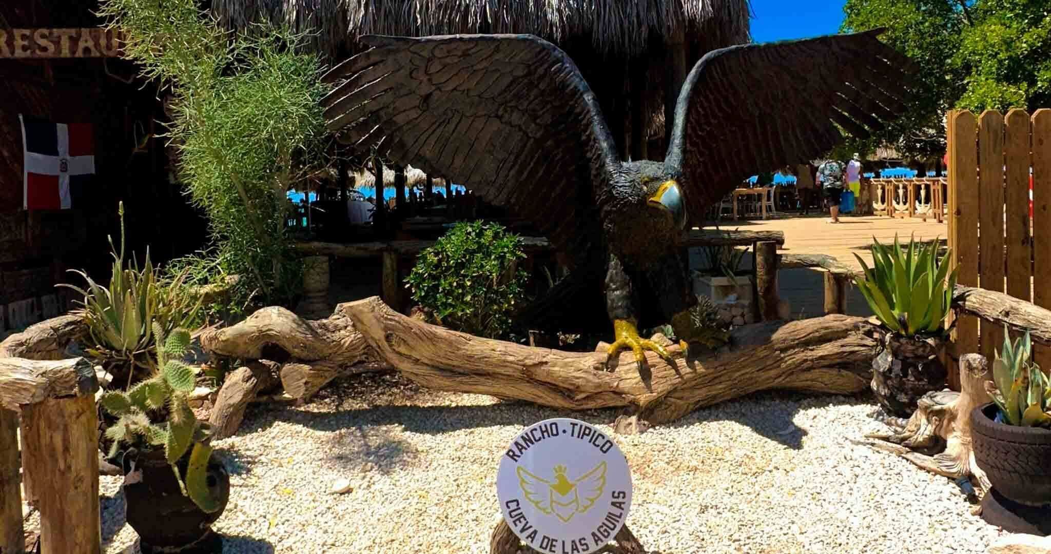 A bird of prey statue in a garden setting with a sign reading "RANCHO TIPICO CUEVA DE LAS AGUILAS".