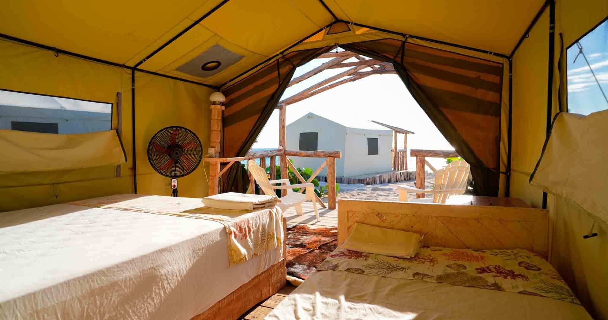 A tented accommodation with two beds, a fan, and a view of the beach and buildings.