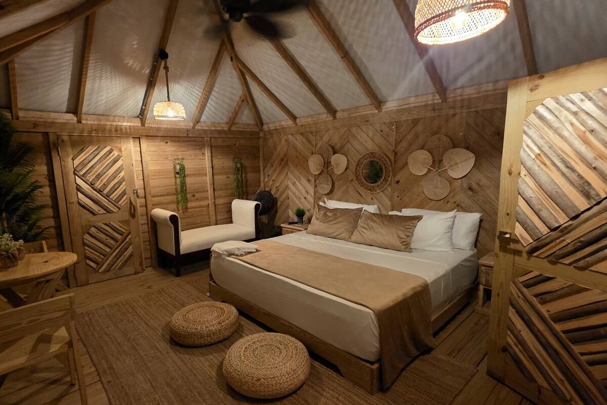 Une pièce chaleureuse, décorée dans un style rustique avec du bois, comprenant un lit, deux chaises, une petite table et un luminaire suspendu.