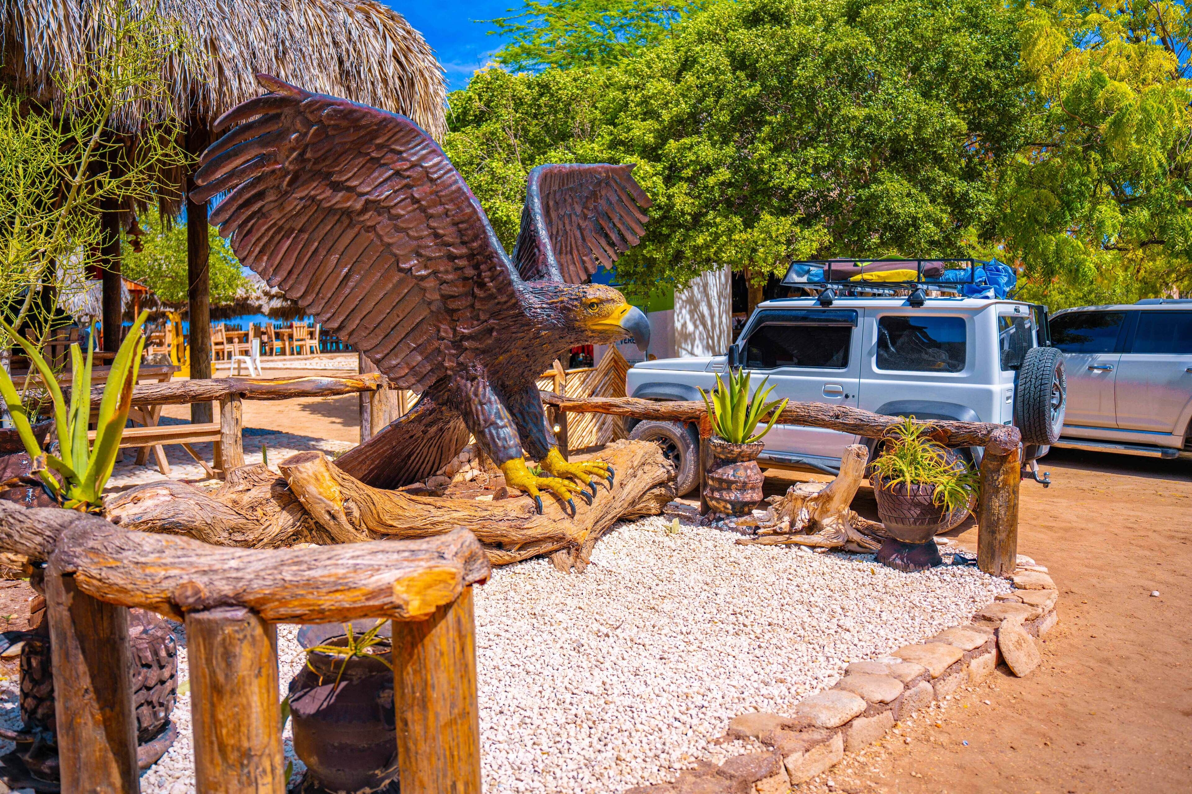 Un espacio al aire libre rústico con una gran escultura de madera de un águila, rodeada de elementos naturales como árboles y una estructura de paja.