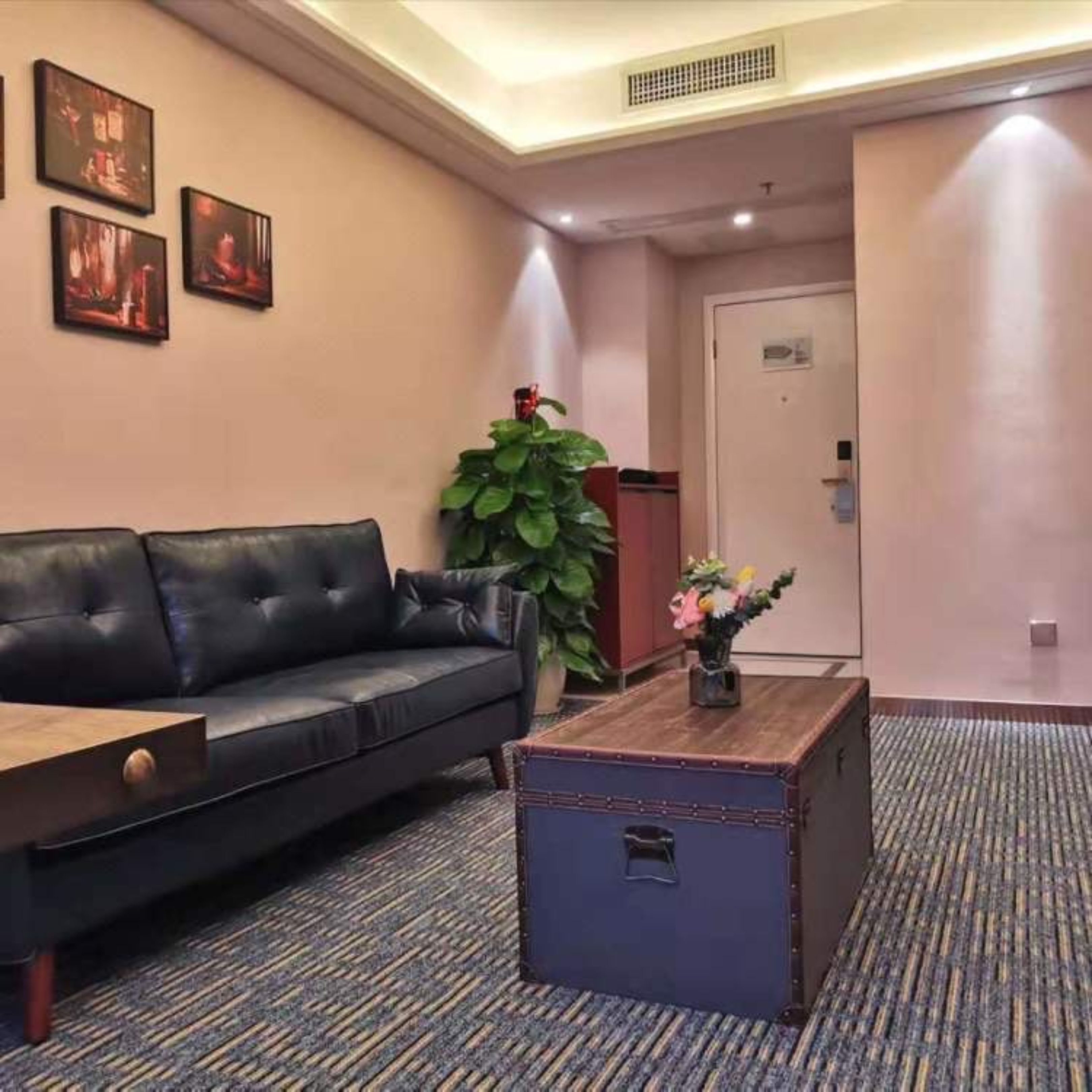 Un hall d’hôtel avec un canapé en cuir noir, une table basse en bois, une plante en pot et une porte visible au fond.