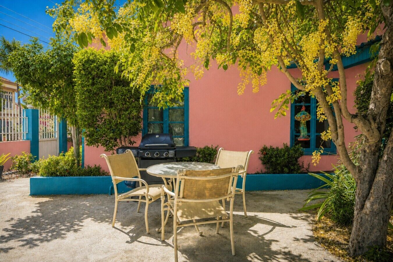An outdoor patio with a round table and two wicker chairs, a grill, and a tree with yellow flowers.