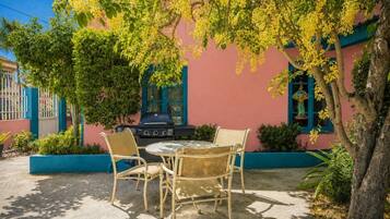 An outdoor patio with a round table and two wicker chairs, a grill, and a tree with yellow flowers.