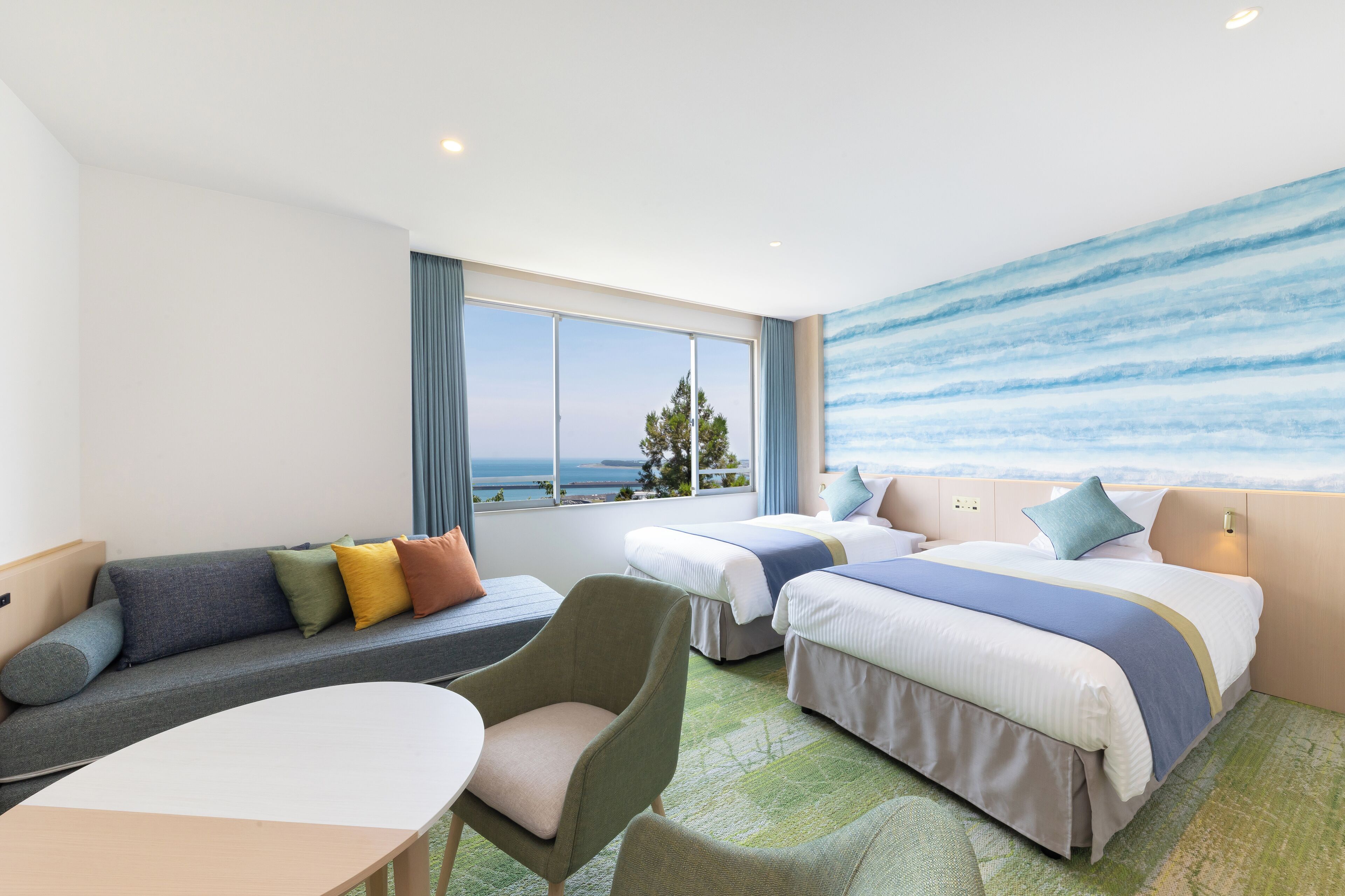 A hotel room with two beds, a sofa, a chair, and a view of the sea.