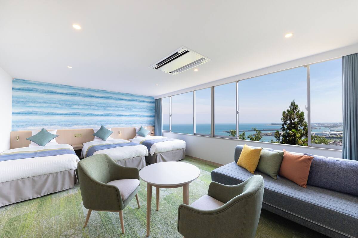 Habitación de hotel con dos camas, un sofá, una mesa redonda y un ventanal con vista al mar.