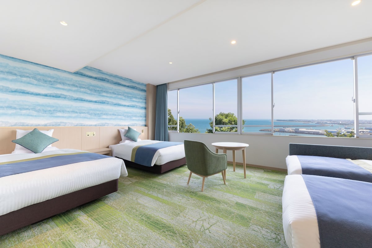 Habitación de hotel con dos camas, una silla verde, una mesita y un amplio ventanal con vistas al mar y al paisaje urbano.