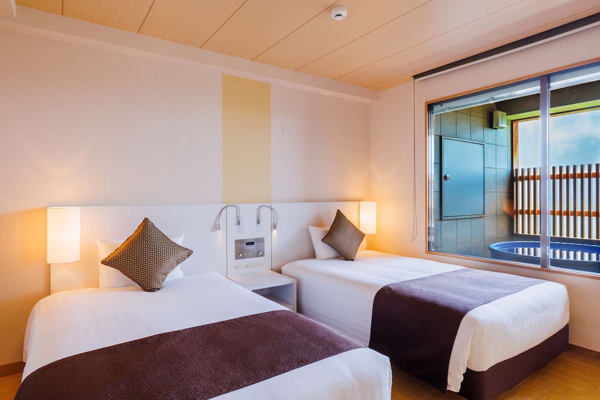 Habitación de hotel con dos camas, una mesita de noche, un panel de control en la pared y una ventana con vistas a un balcón.