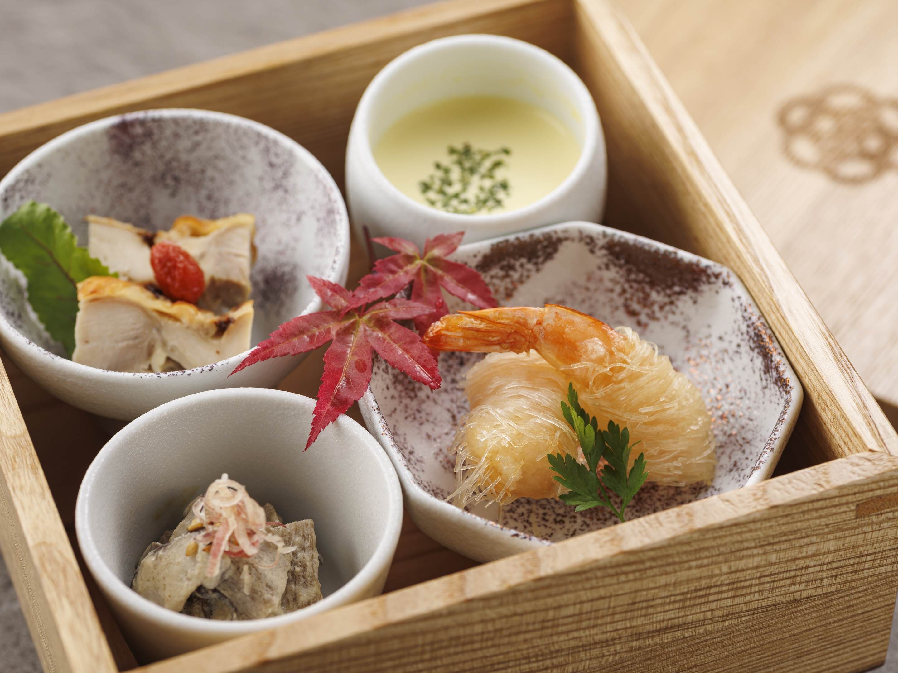 A wooden tray with four bowls containing various food items, including a shrimp and some leafy greens.