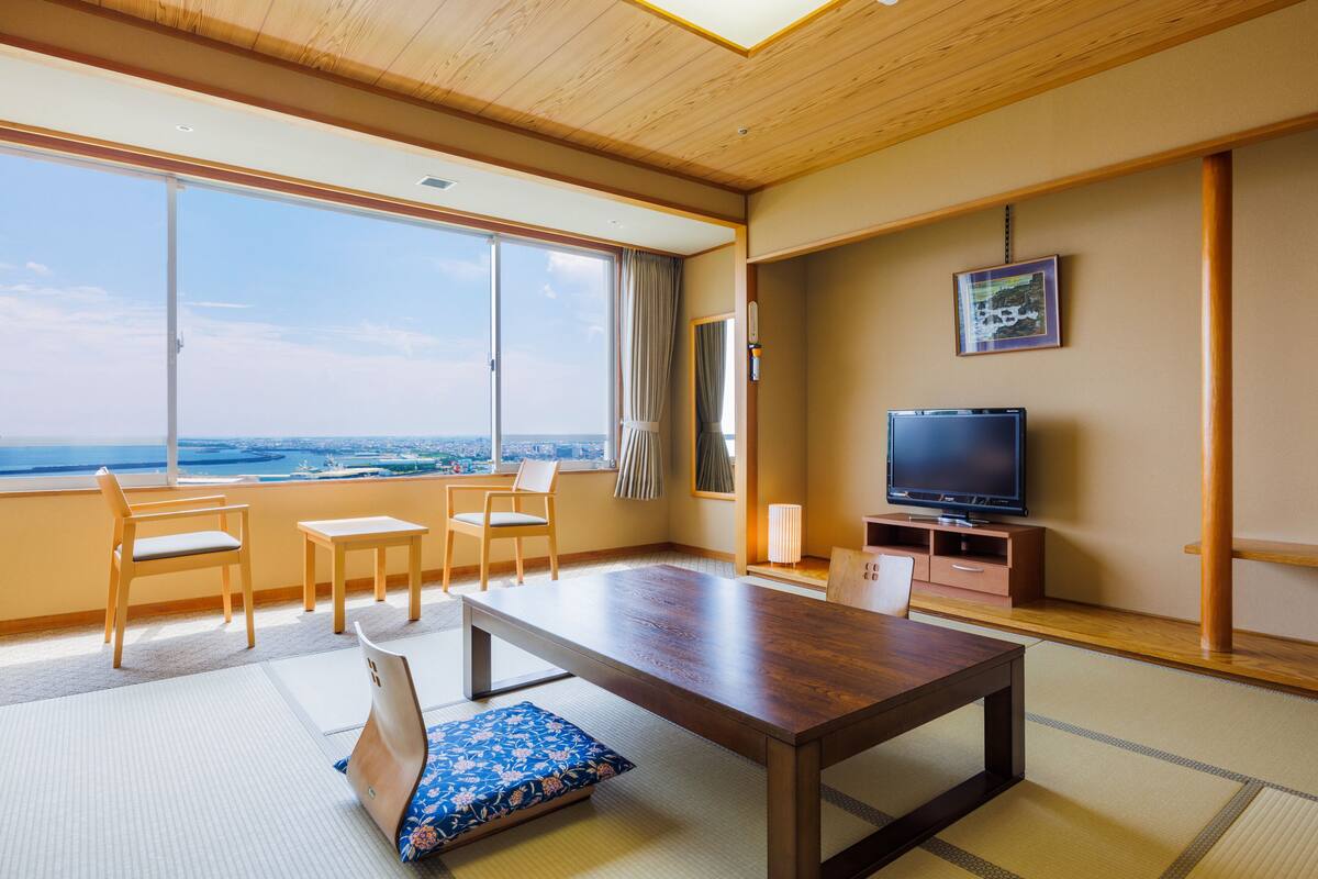 Habitación de hotel con un ventanal grande, techo de madera, televisor, mesa de comedor y un sofá con cojín.