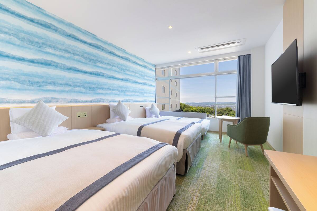 Habitación de hotel con dos camas, un escritorio, una silla, un ventanal grande y un mural en la pared.