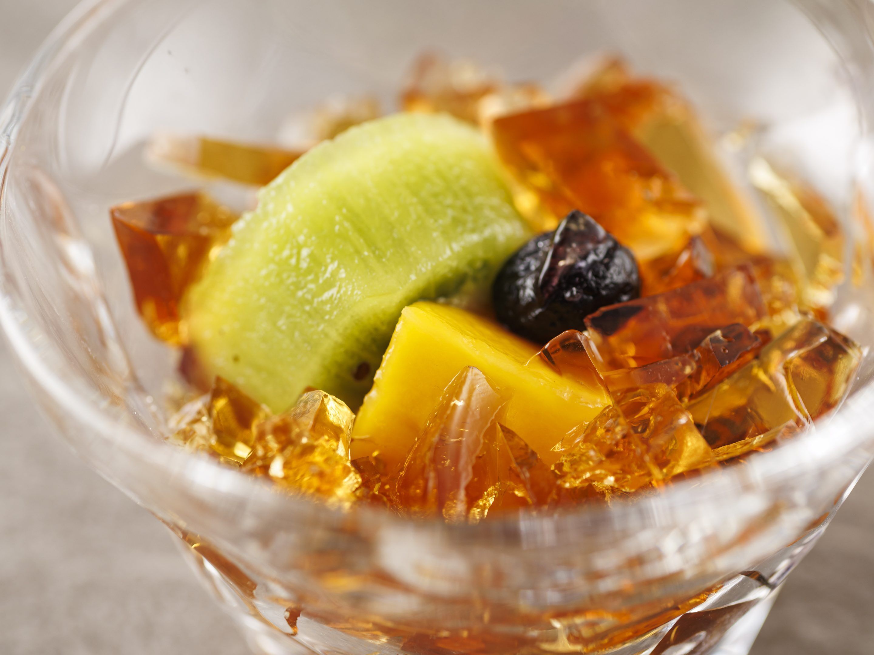 A clear glass bowl containing a dessert with a green scoop, yellow cubes, and a dark fruit.