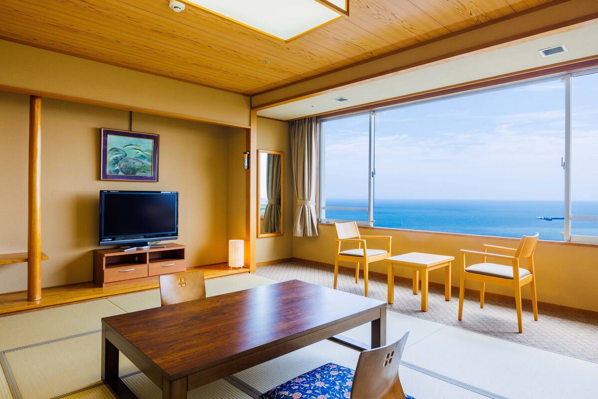 Habitación de hotel con techo de madera, televisor sobre un soporte de madera, ventana grande con vistas al mar y suelo con tatami japonés tradicional.