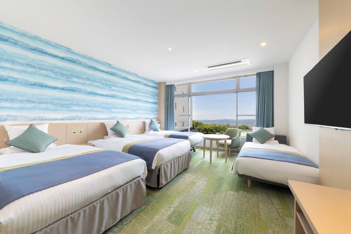Habitación de hotel con tres camas, una mesita pequeña y un ventanal con vistas.