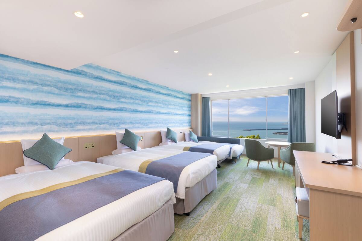 Habitación de hotel con tres camas, un escritorio, un televisor y un ventanal con vistas al mar.