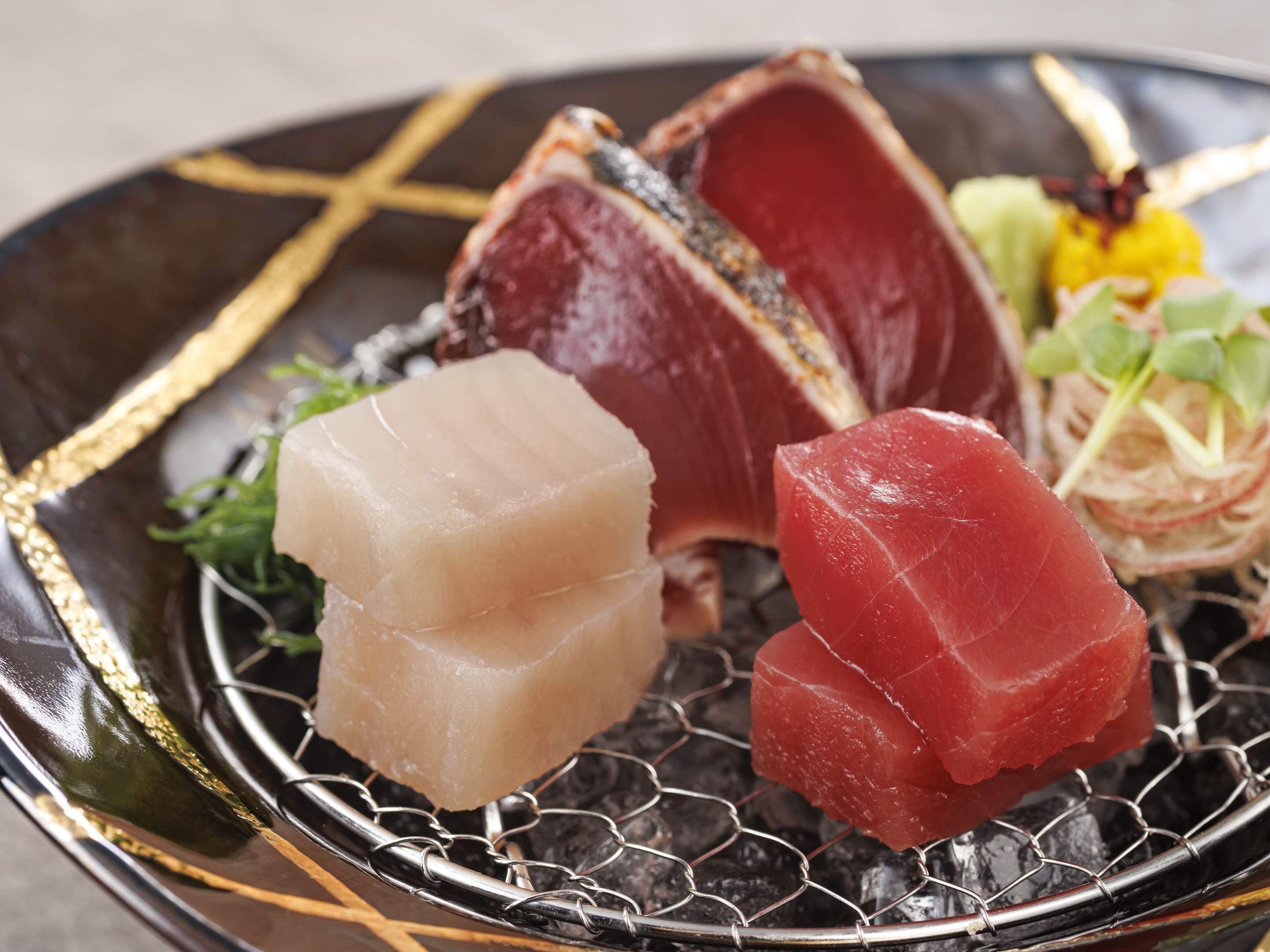 A plate of sashimi with various cuts of fish, including a piece of salmon and tuna, served on a black plate with a gold design.