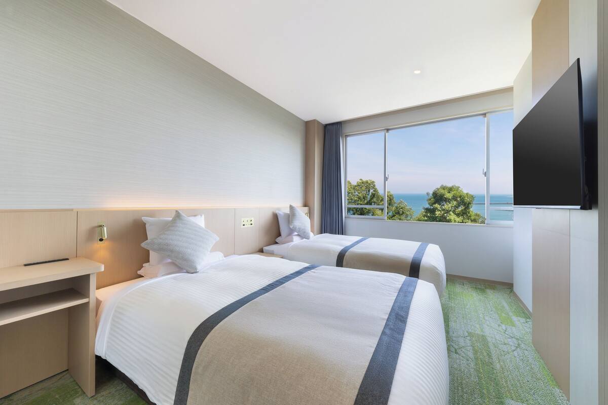 Habitación de hotel con dos camas, un escritorio, un televisor de pantalla plana y un amplio ventanal con vistas al mar.