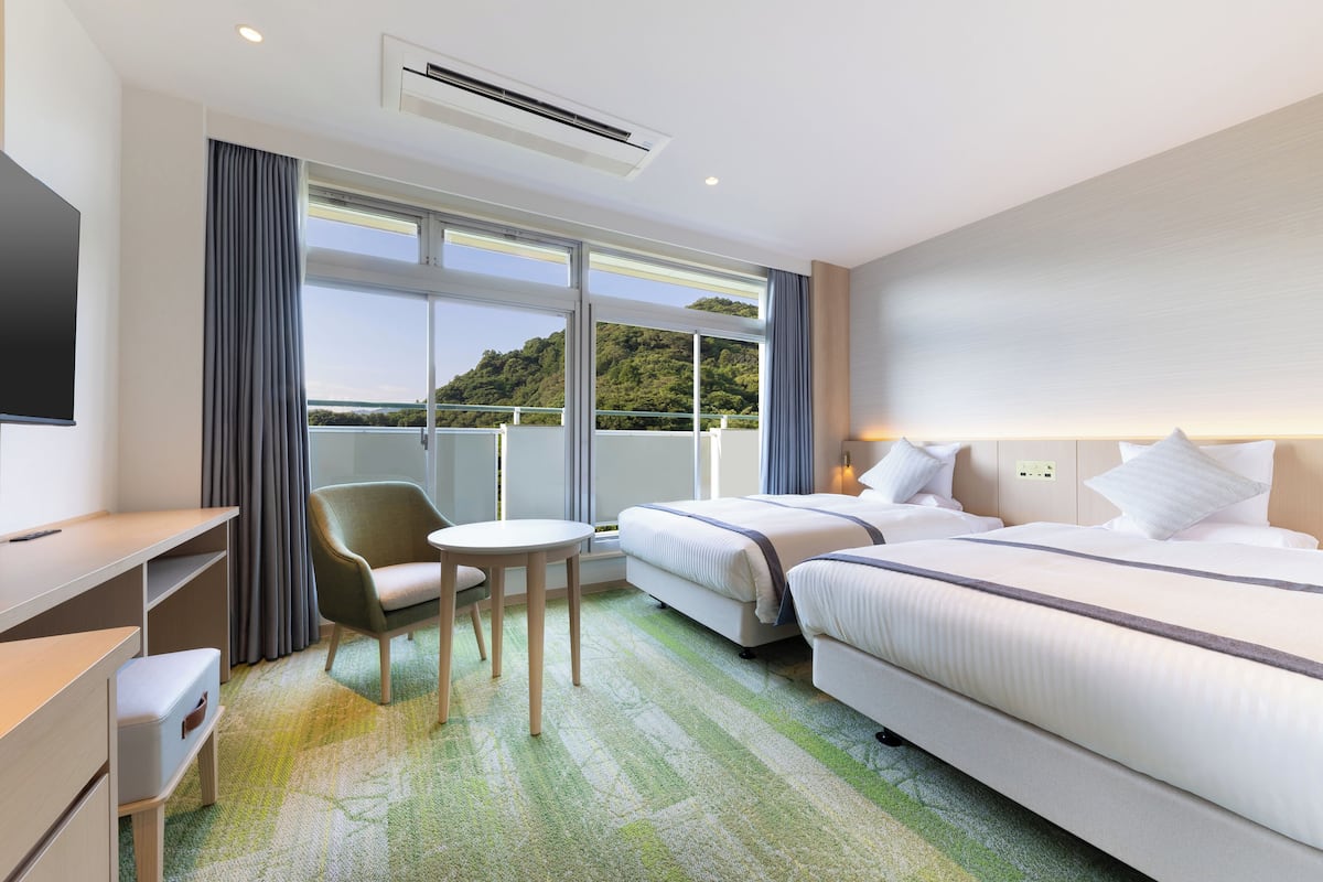 Habitación de hotel con dos camas, un escritorio, una silla y un amplio ventanal con vistas a un paisaje verde.