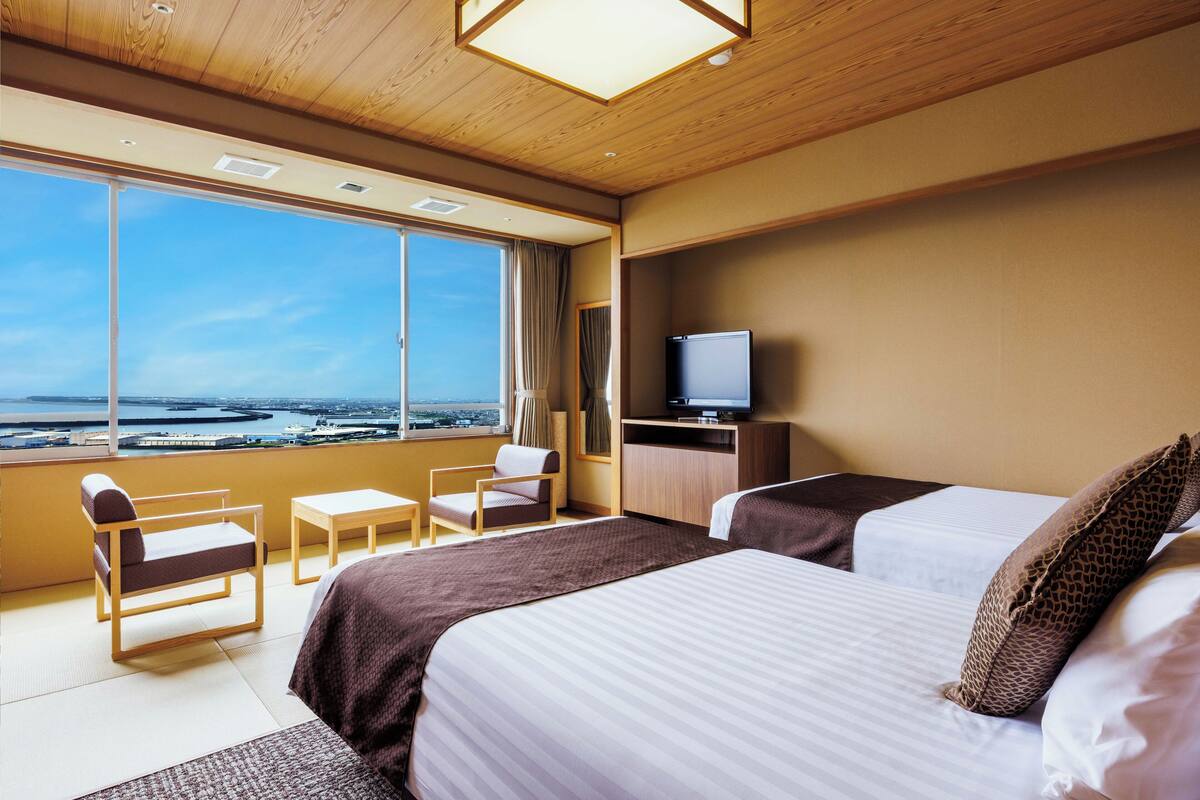 Habitación de hotel con dos camas, un escritorio, una silla, televisión y un ventanal con vista al mar.