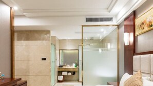 A hotel room with a large bed, a wooden dresser, a vanity with a mirror, and a bathroom visible through a glass partition.