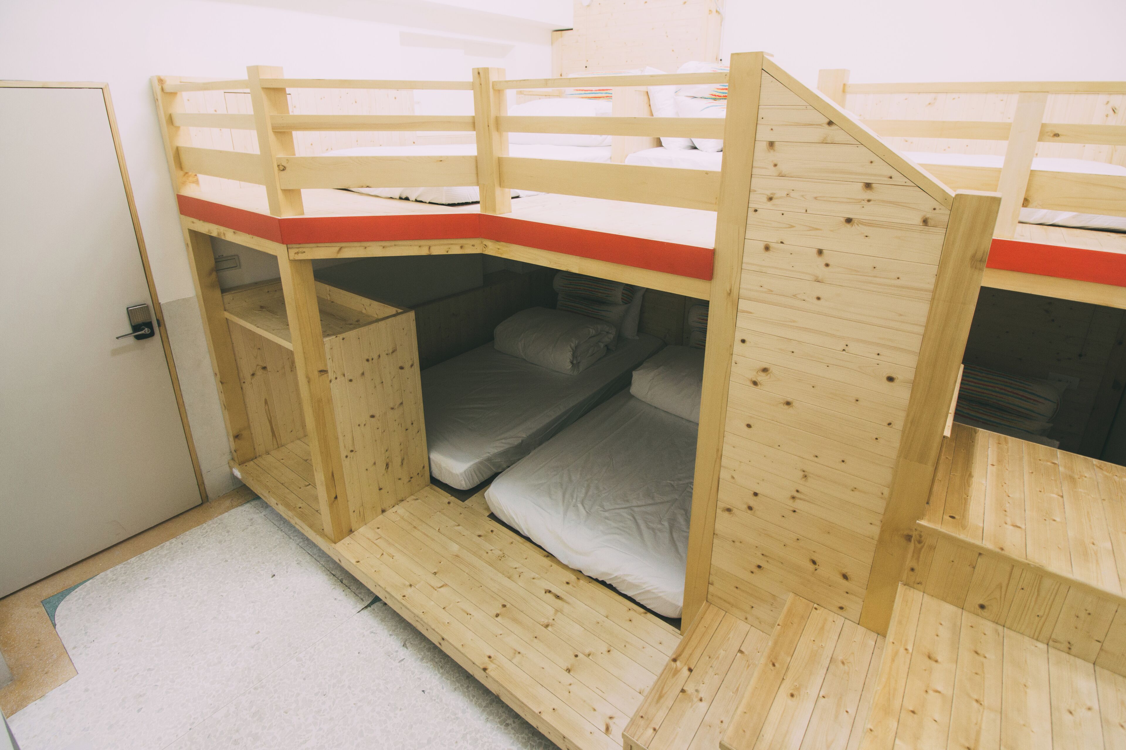A bunk bed room with a wooden frame, a red mat at the top, and a door on the left.
