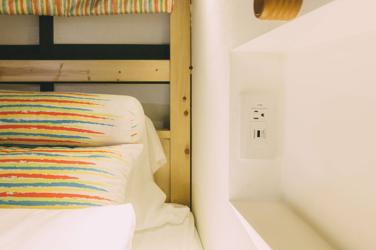 Ein Stockbett mit farbig gestreifter Bettwäsche, einem Holz-Kopfteil und einer weißen Wand mit Steckdose.