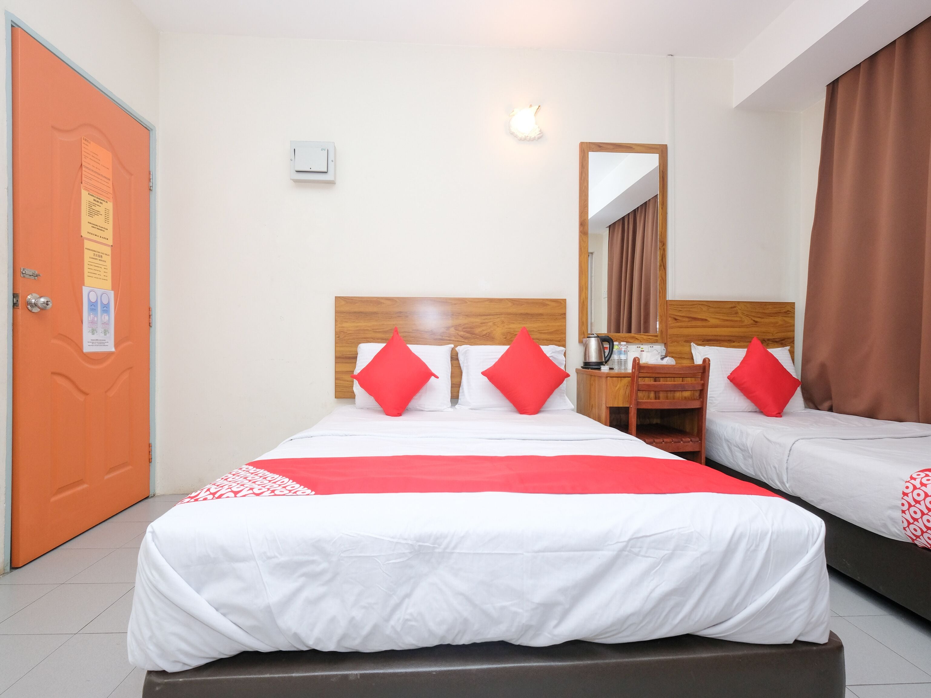 A hotel room with two beds, a wooden headboard, a mirror, a desk with a kettle, and a brown curtain.