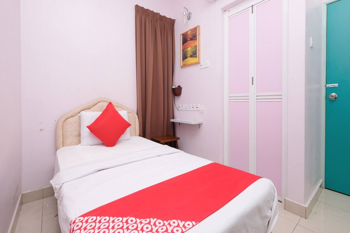 Ein Schlafzimmer mit einem Bett, einem roten Kopfkissen, einer weißen Bettdecke, einem braunen Vorhang, einer Wandleuchte, einem Gemälde, einer türkisblauen Tür und einem weißen Nachttisch.