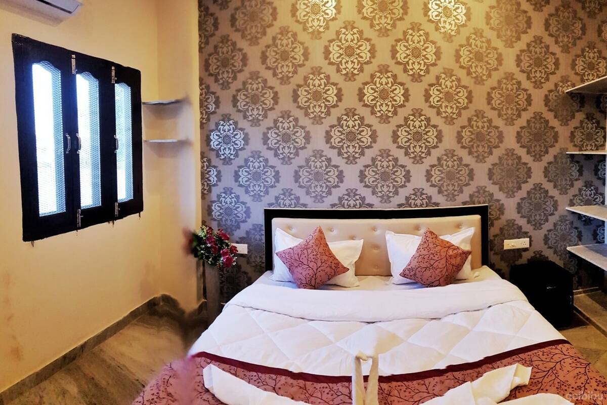 A hotel room with a bed, a window with blinds, a patterned wall, and a small desk with a chair.