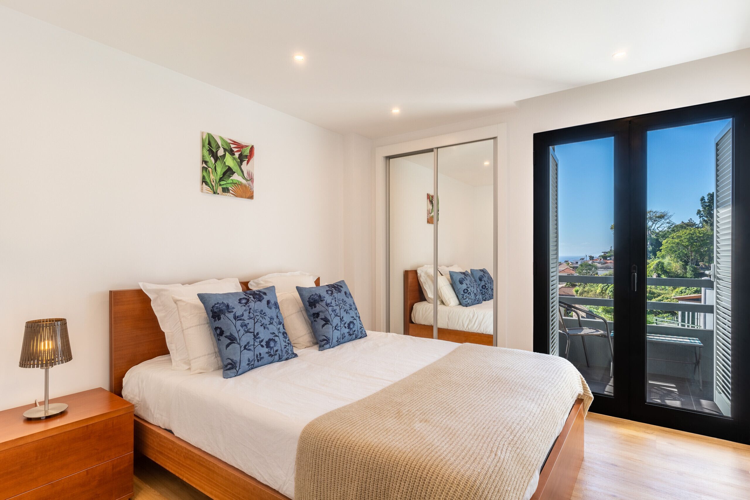 A bedroom with a large bed, a nightstand, a mirror, and a view of the outdoors through a sliding door.