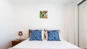 Ein ordentlich bezogenes Bett mit weißer Bettwäsche und zwei blau blumigen Kissen, ein kleiner Nachttisch mit Lampe sowie ein farbenfrohes abstraktes Gemälde an der Wand.