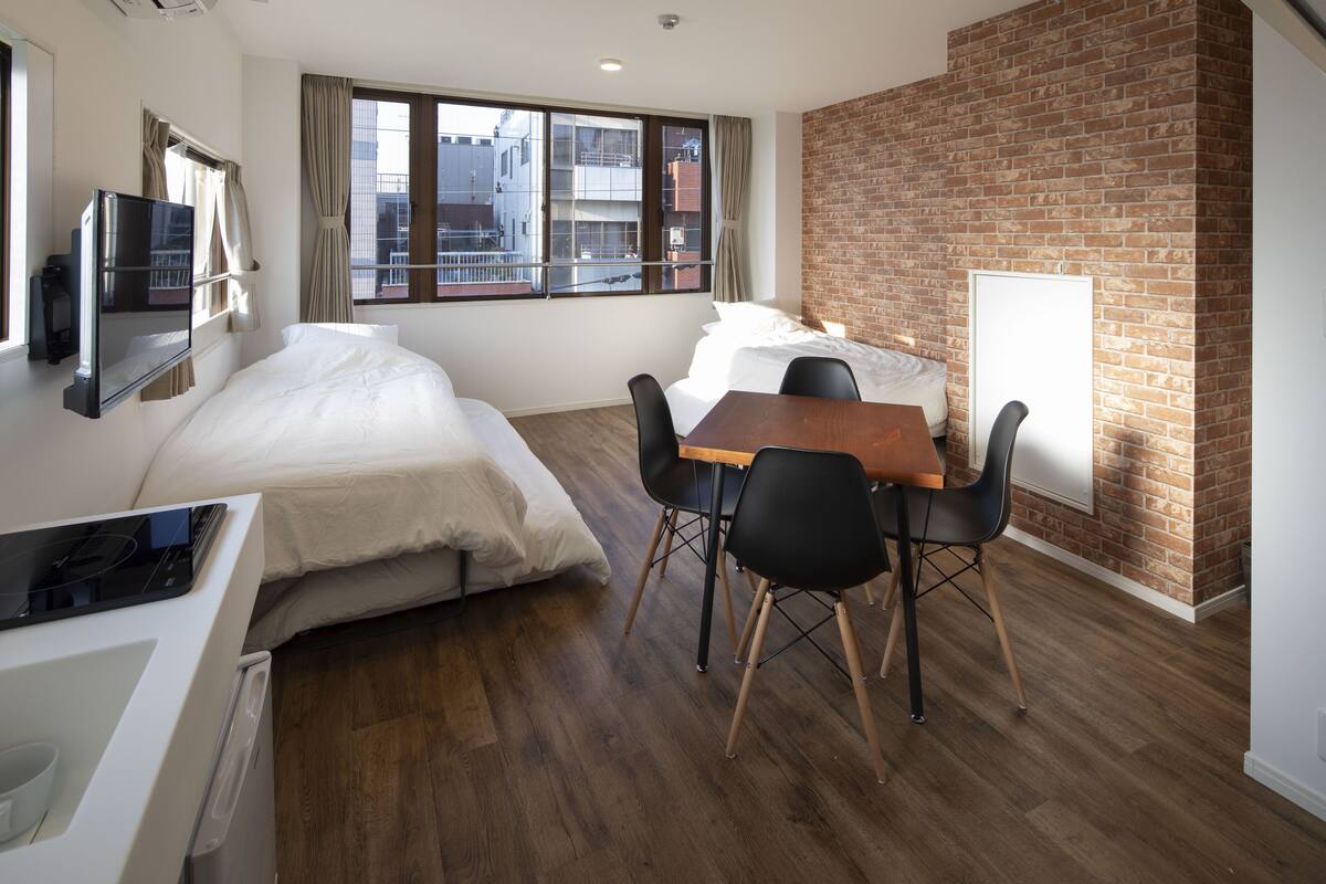 Une chambre d’hôtel moderne avec un mur d’accent en briques, une grande fenêtre donnant sur la ville, un lit, un bureau avec une télévision et un coin repas avec des chaises.
