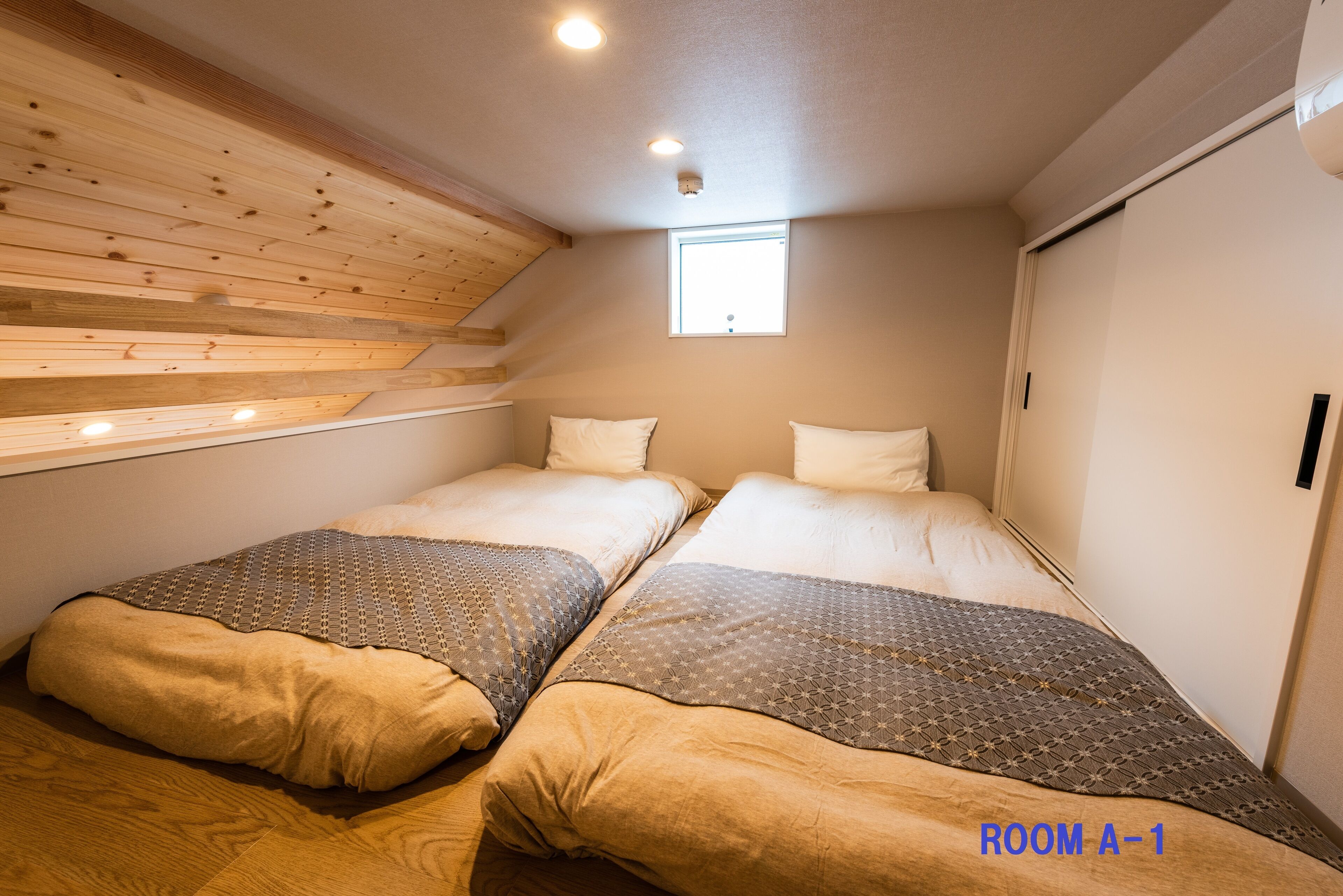 A bedroom with two beds, wooden slats on the ceiling, a small window, and a door.
