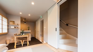 Una habitación con suelo de tatami, una mesa de madera con sillas, un estante con objetos decorativos y una escalera.