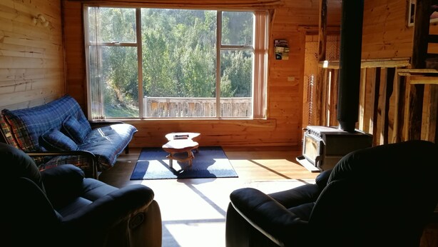 A cozy cabin interior with a large window, wooden walls, a blue sofa, two chairs, and a fireplace. - Minnow cabins, peaceful bush setting, cabin 2 (Lower Beulah)