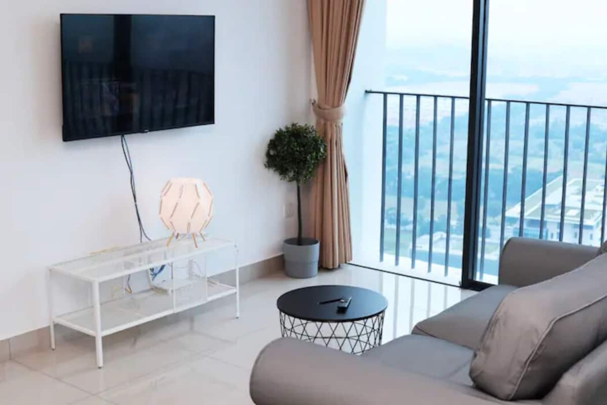 Un salon moderne avec un téléviseur à écran plat fixé au mur, une table basse en verre, un canapé gris et une vue sur la ville grâce à de grandes fenêtres.