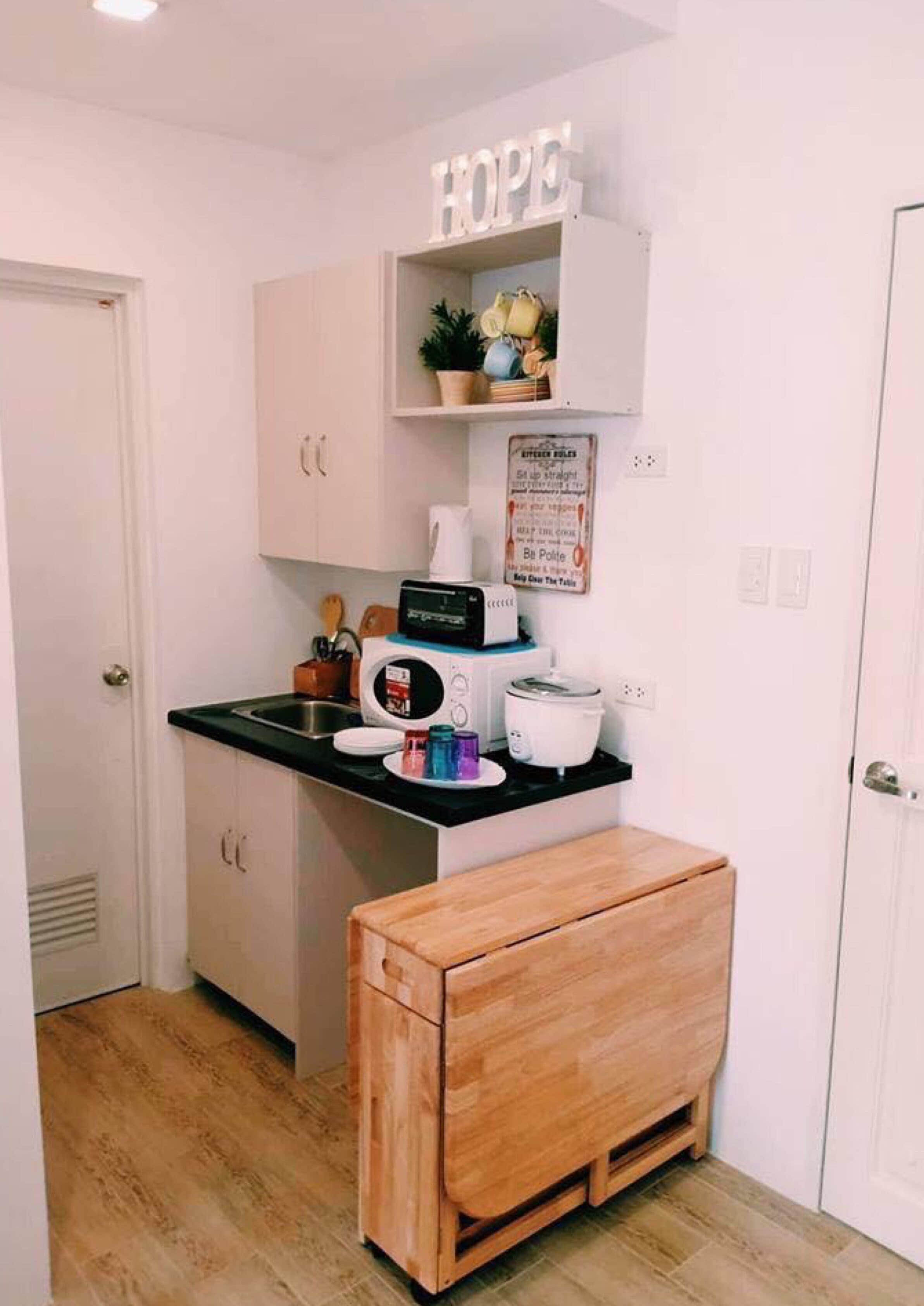 Cocina compacta con armarios blancos, encimera negra, microondas, tostadora y un banco de madera.