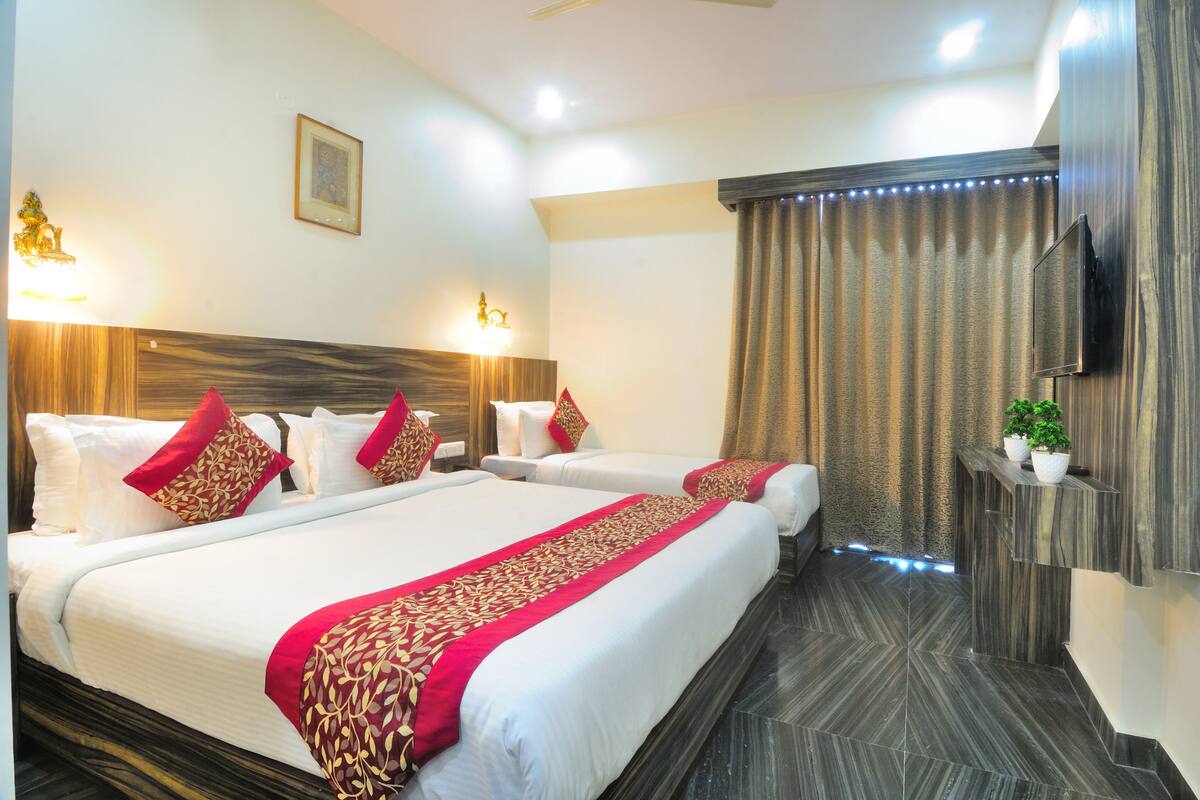 A hotel room with two beds, each with white linens and red accents, a flat-screen TV mounted on the wall, and a small plant on a shelf.