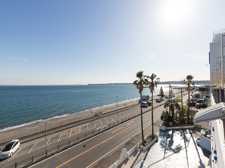 A coastal road with a beach and ocean on one side, and a parking area with palm trees and a building on the other.
