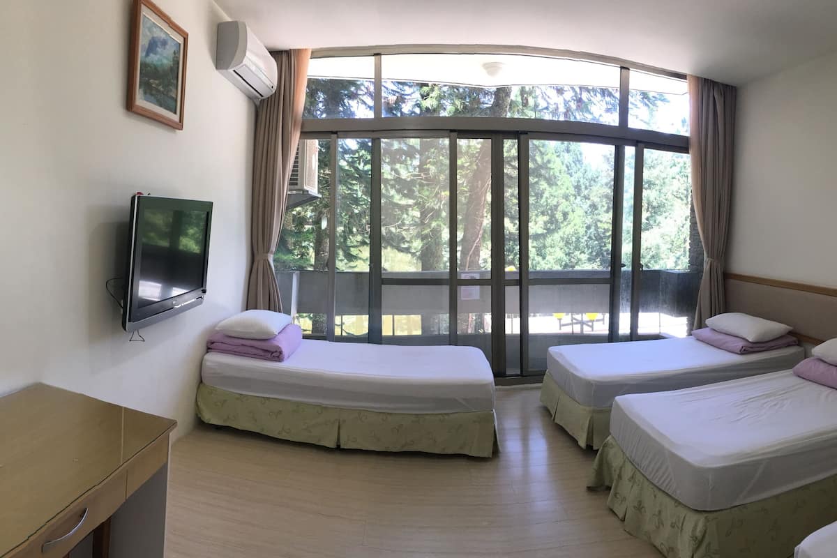 Ein Hotelzimmer mit zwei Betten, einem Fernseher, einem Schreibtisch und einem Fenster mit Blick auf Bäume.
