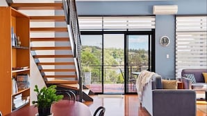 A modern living room with a wooden staircase, a sofa, a dining table, and a view of greenery through large windows. - Cove View Retreat (St Helens)