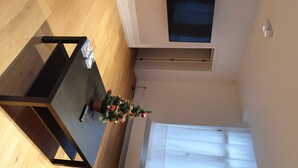 Habitació amb terra de fusta, taula de dinar amb un arbre de Nadal, televisió penjada a la paret i finestra amb persianes.