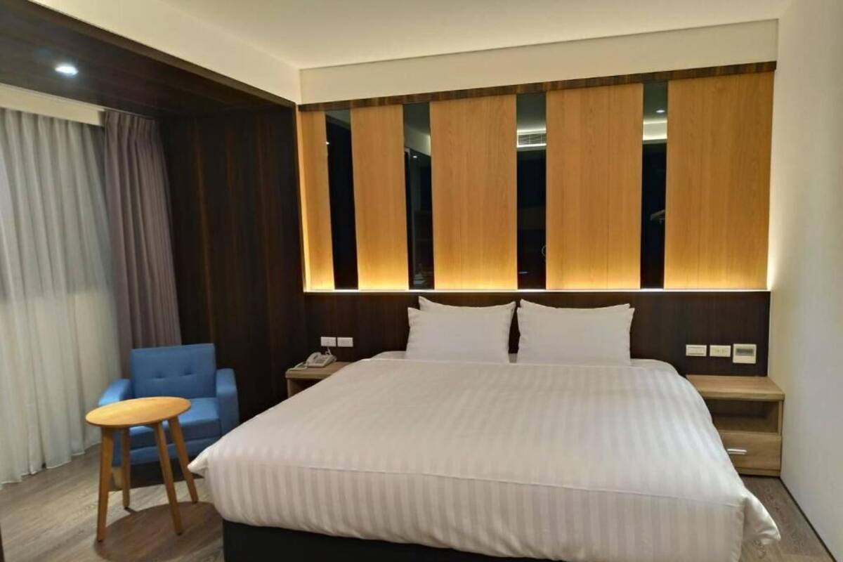 Une chambre d’hôtel avec un grand lit, une tête de lit en bois, une chaise bleue et un petit tabouret en bois.