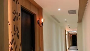 Un long couloir étroit, avec une porte en bois sur le côté gauche, un luminaire au-dessus et une grille de ventilation au plafond.