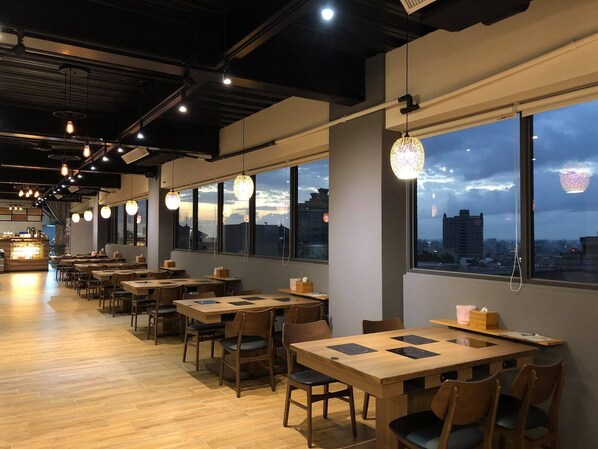 Un restaurante moderno con mesas y sillas de madera, ventanas amplias con vistas a la ciudad e iluminación colgante.